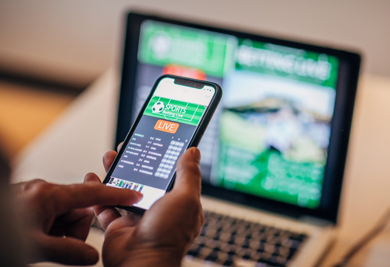 Hand holding a smartphone that shows a gambling app. A laptop in the background is also on a gambling site.